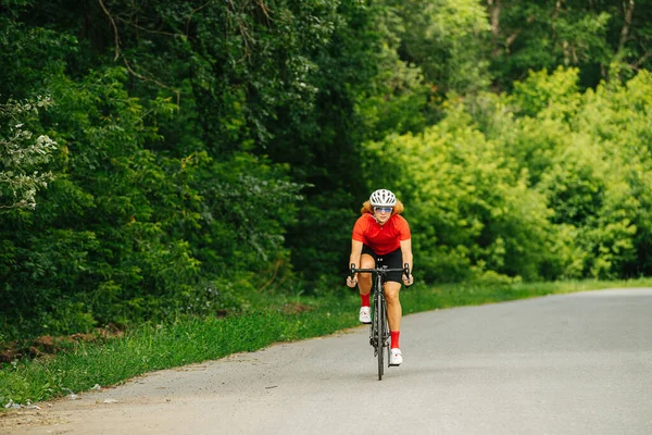 Athletic woman in a professional gear on a slim bike in a white perforated helmet. Riding on a road with trees on sides.