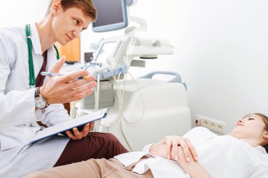 Genç erkek doktor, ultrason teşhis kabinesinde muayene koltuğunda genç bayan hastayla sohbet ediyor. Not alıyor, hastanın geçmişini dolduruyor..