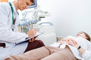 Genç erkek doktor, ultrason teşhis kabinesinde muayene koltuğunda genç bayan hastayla sohbet ediyor. Not alıyor, hastanın geçmişini dolduruyor..