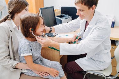 Muayenede annesinin kucağında oturan küçük kız. Doktor çocuk doktoru. Steteskopla oynayıp güvenli olduğunu gösterelim..
