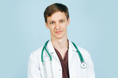 Mavi üzerine beyaz bornoz ve kahverengi gömlek giymiş genç bir erkek doktorun portresi.