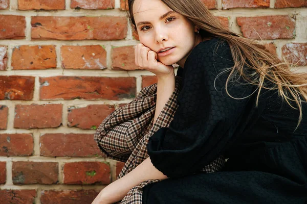 Side portrait of a crouching young woman in a long black dress in front of a old house brick wall. Holding checkered jacket. Cropped at the top.