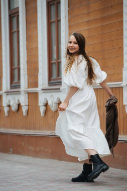 Beyaz pofuduk bir elbise ve ağır çizmelerle asfalt yolda yürüyen mutlu genç bir kadın. Büyük ahşap bina boyunca yürüyor. Elinde kareli ceket taşıyor. Kameraya bakıyorum..