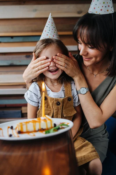 Mother and daughter celebrating birthday in a cafe. Mom covering kid\'s eyes, surprising her with a cake and lighted candle. Both wearing party cone hats.