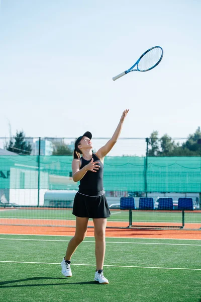 新しいテニスコートで黒のスポーツ衣装のトレーニングで10代の女の子は ラケットで遊んで 空気中でそれを投げます フェンスや高層ビルを覆う旗の上に — ストック写真