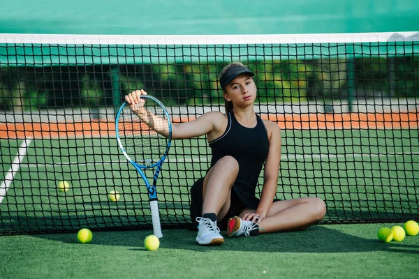 晴れた日に真新しいテニスコートの真ん中に座っているスポーツ服の10代の少女 彼女はトップとキャップ付きの短いテニススカートを着ている ネット上でのリース — ストック写真