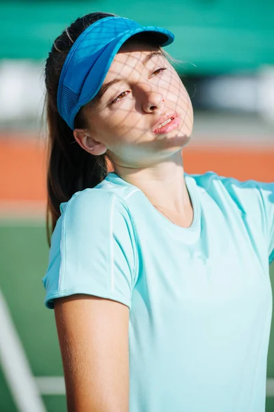 ラケットから顔にネットの影を持つ青いテニスキャップの10代の女の子の肖像画 彼女は顔に顔をしかめっ面で — ストック写真