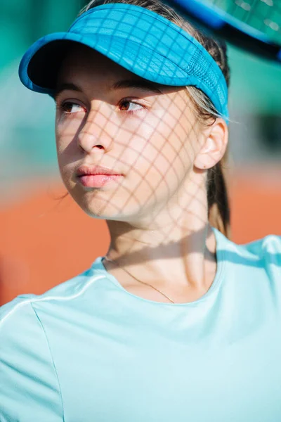 ラケットから顔にネットの影が付いている青いテニスキャップのきれいな10代の女の子の肖像画 彼女はリラックスして横を見て — ストック写真
