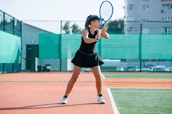 黒のスポーツ衣装を着た10代の少女が新しいコートでテニスをし ボールを速く返します フェンスや高層階のアパートの建物をカバーするバナーの上 サイドビュー — ストック写真