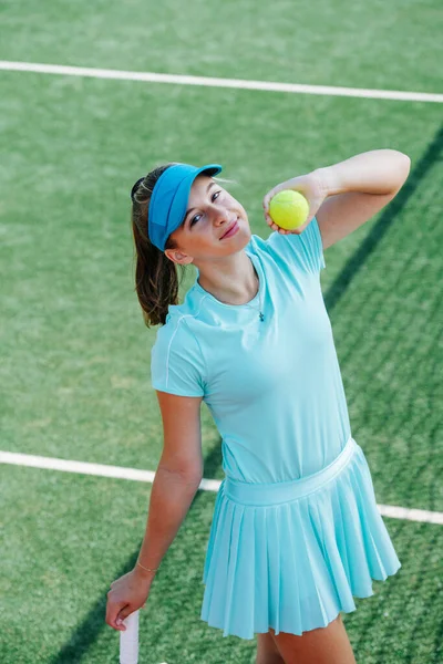 Teenage girl in a sky blue sportive outfit posing with ball in her hand on a tennis court. High angle. Looking at the camera.