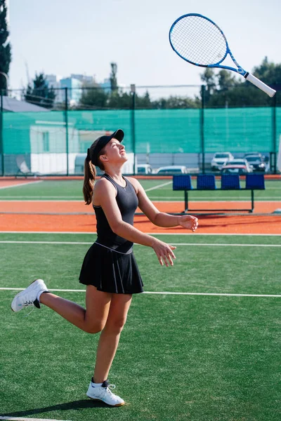 新しいテニスコートで黒のスポーツ衣装のトレーニングで10代の女の子は ラケットで遊んで 空気中でそれを裏返します フェンスや高層ビルをカバーするバナーの上に サイドビュー — ストック写真