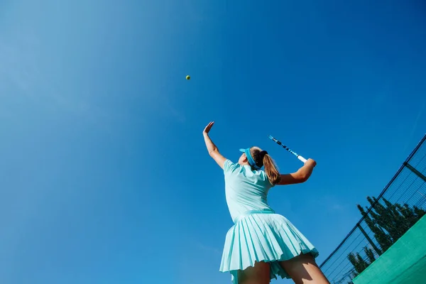 空の青のスポーツ衣装を着た10代の女の子がテニスをし ボールを空中に投げ 奉仕を練習します 低い角度だ 深い青空の上に — ストック写真