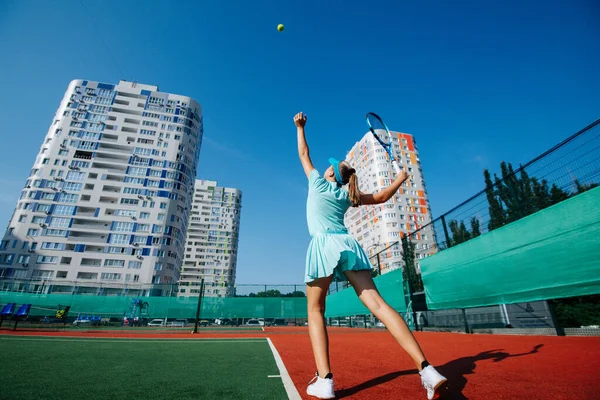 新しいテニスコートで空の青のスポーツ衣装の訓練で10代の女の子は 奉仕を練習し 空気中のボールを投げます 低い角度だ 空と高層階建ての新しいカラフルな建物の上に — ストック写真