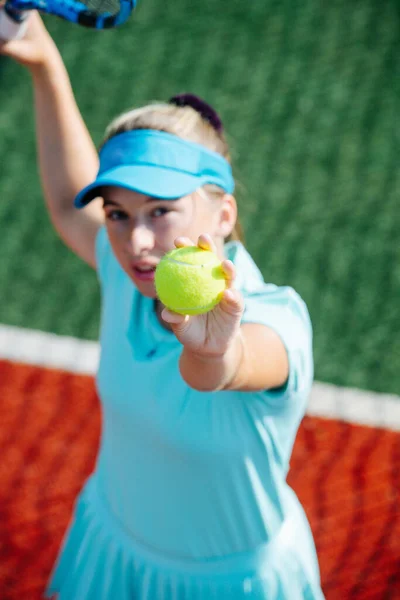 新しいテニスコートで空の青のスポーツ衣装の訓練で10代の女の子は 奉仕を練習し 空気中のボールを投げます 高い角度だ カメラを見ると — ストック写真