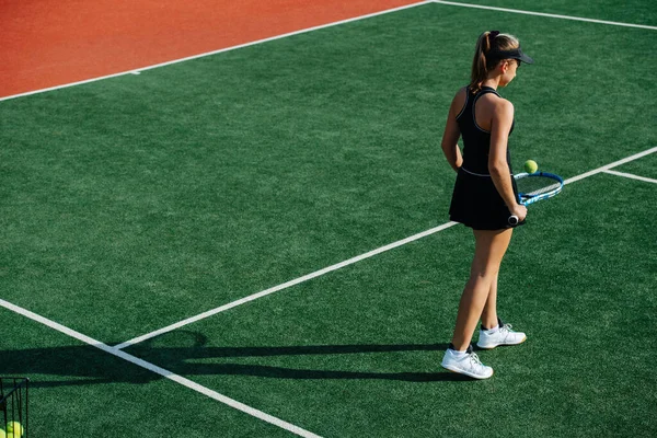 晴れた日に真新しいテニスコートの真ん中に立っているスポーツ衣装を着た10代の少女 彼女はトップとキャップ付きの短いテニススカートを着ている 彼女のラケットからボールをバウンス 後ろからの眺め — ストック写真