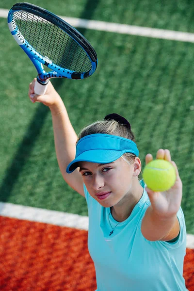 新しいテニスコートで空の青のスポーツ衣装の訓練で10代の女の子は 奉仕を練習し 空気中のボールを投げます 高い角度だ カメラを見ると — ストック写真