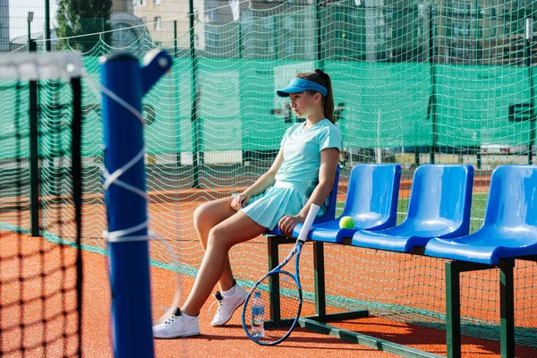 短い休憩を取るためにテニスコートの横にあるプラスチック製の椅子ベンチに座って空の青のスポーツ衣装を着た10代の女の子 現場を見てる サイドビュー — ストック写真