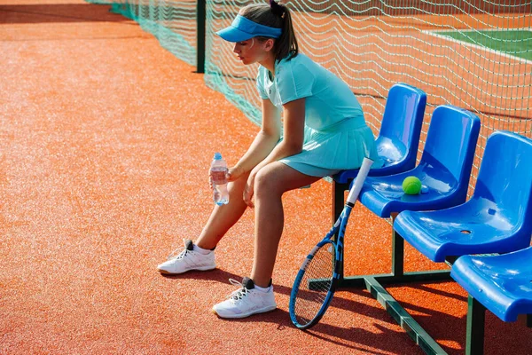 テニスコートの横にあるプラスチック製の椅子ベンチに座って短い休憩を取るために空の青のスポーツ衣装を着た思慮深い10代の女の子 思考では 水のボトルを保持失われます サイドビュー — ストック写真
