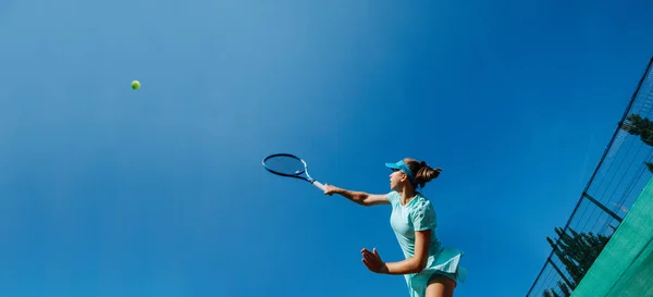 空の青のスポーツ衣装を着た10代の女の子がテニスをして ラケットでボールを高く上げています 低い角度だ 深い青空の上に — ストック写真