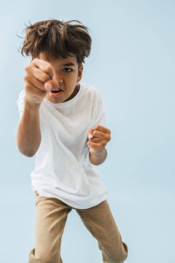 Hintli Asyalı çocuk kamerayı kötü kapalı yumruklarıyla dürtüyor, yumruk atıyor gibi yapıyor. Kameraya bakıyorum. Mavi arkaplan.