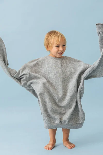 Αστεία μικρό αγόρι αιωρείται μανίκια από τεράστια υπερμεγέθη γκρι παχύ ζεστό πουκάμισο — Φωτογραφία Αρχείου