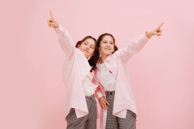 Güzel küçük ikiz kardeşler mutlu bir şekilde gülümsüyor, farklı yönleri gösteriyorlar. Pembe arka planda, stüdyo çekimleri. Birbirlerine yakın dururlar. İşaret ediyor, bakıyor ve uzaklarda.