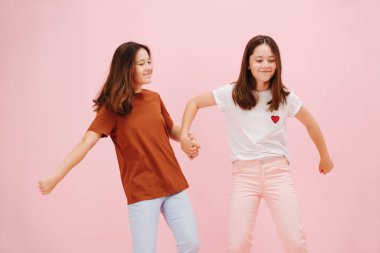 Güzel küçük ikiz kardeşler mutlu bir şekilde el ele tutuşuyorlar. Pembe arka planda, stüdyo çekimleri. Onlar gömleklerinin aksine pembe ve mavi pantolon giyerler. Bir kız diğerini çekiyor..