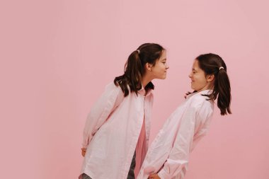 Güzel küçük ikiz kardeşler mutlu bir şekilde gülümsüyor, farklı yönlere bakıyor. Pembe arka planda, stüdyo çekimleri. Biri ciddi yüzünü yakınlaştırıyor, diğeri ise kapalı gözlerle kaçıyor..
