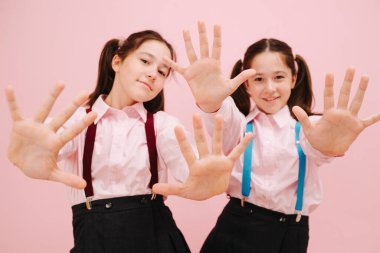 İkiz okullu güzel kızlar kameraya el kaldırıyor. Düşük açı. Pembe arka planda, stüdyo çekimleri. Eteklerinde farklı renkte diş telleri var..