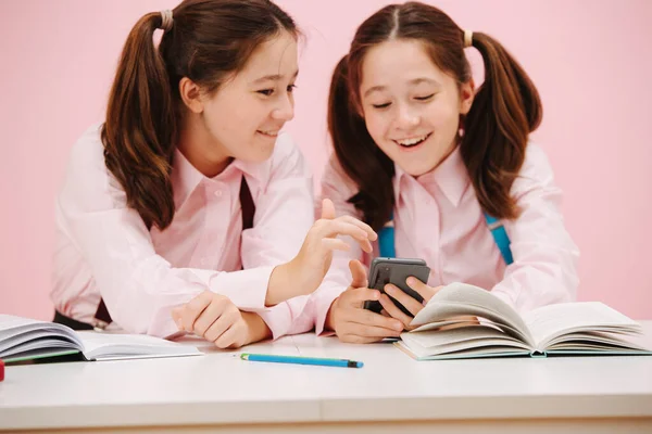 漂亮的双胞胎女学生坐在课桌后面 前面的观点 粉红背景 工作室拍摄 他们厌烦了 分享智能手机的形象 — 图库照片