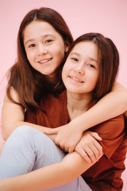 Güzel ikiz kız kardeşlerin mutlu bir şekilde kameraya baktıkları portre. Pembe arka planda, stüdyo çekimleri. Yerde oturuyor, biri diğerine sarılıyor..