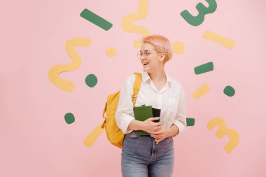 Kısa boyalı sarı saçlı ve yuvarlak gözlüklü genç bir kadın. Anne kotu, gömlek ve sarı sırt çantası giyiyor. Not defterlerini tutuyorum. Üzerine yeşil ve sarı kağıt şekiller yapıştırılmış pembe duvarlar..