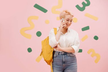 Kısa boyalı sarı saçlı ve yuvarlak gözlüklü genç bir kadın telefonda konuşuyor. Anne kotu, gömlek ve sarı sırt çantası giyiyor. Üzerine yeşil ve sarı kağıt şekiller yapıştırılmış pembe duvarlar..