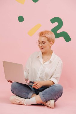 Yuvarlak gözlüklü, kısa sarı saçlı, dizüstü bilgisayarla bağdaş kurmuş genç ve mutlu bir kadın. Üzerine yeşil ve sarı kağıt şekiller yapıştırılmış pembe duvarlar..