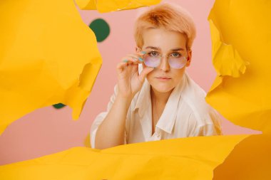 Geniş bir delikten bakan kuşkucu genç bir kadın, büyük, sıkışık sarı bir kağıt parçasına yırtılmış. Kısa sarı saçları, yuvarlak gözlükleri ve yıldız makyajı var..