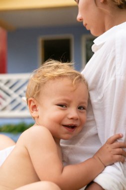 Annesinin omzuna sarılan mutlu, sarışın, yeni yürümeye başlayan bebek. Akıllı telefonuyla sosyal ağlarda çok meşgul. Salon sandalyesi, tatil evinin önündeki güneşli teras..