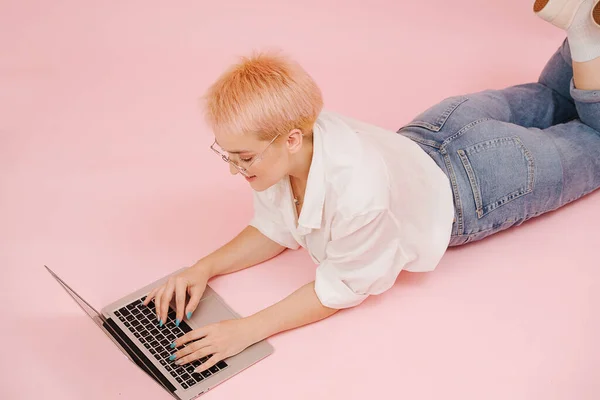 短い金髪の髪をした若い女性が床に横たわって ラップトップで作業しています 彼女は母のジーンズとドレスシャツを着ている ピンクの壁に緑と黄色の紙の形が接着されています — ストック写真