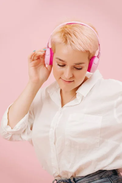 Immersed young woman with short hair enjoying music, listening to wireless headphones. Over pink background. She has short dyed blond hair and stars eye makeup. Looking down with smile.