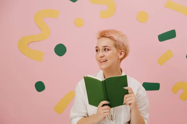 緑の表紙の本を手にした幸せな若い女性 ブロンドの髪に星のメイクを施しています ピンクを背景に — ストック写真