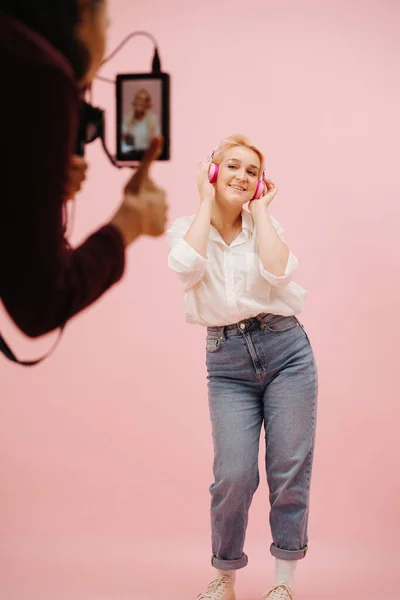 フィールドモニターを使用して ワイヤレスヘッドフォンでポーズ若い女性を撮影女性カメラマン ピンクを背景に モデルは短めのブロンドの髪を染め 母のジーンズとドレスシャツを着ています モデルに焦点を当てる — ストック写真