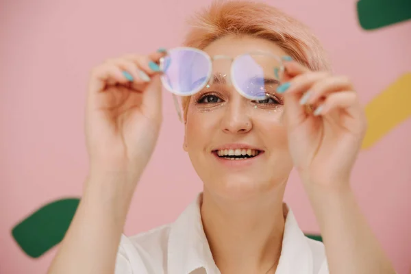 Happy young woman looking at glasses she\'s holding. She has short dyed blond hair and round glasses and stars eye makeup. Over pink background. Mouth open in a smile.