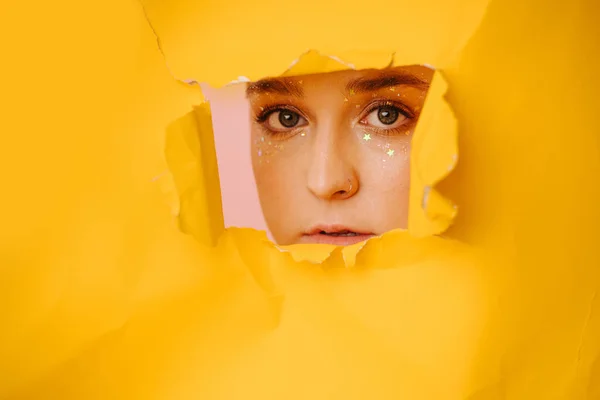 Young woman peeking through a hole in a big sheet of cramped yellow paper. She has short dyed blond hair and stars eye makeup.