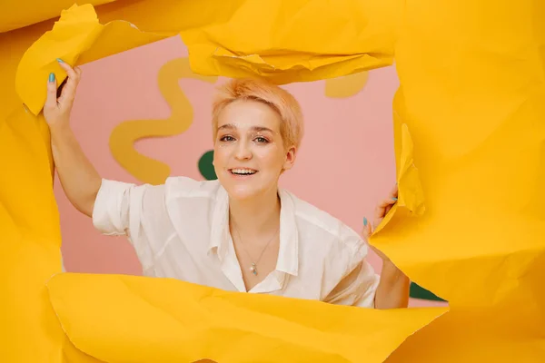 Smiling young woman emerging through a hole in a big sheet of cramped yellow paper, tearing it up. She has short dyed blond hair and stars eye makeup.