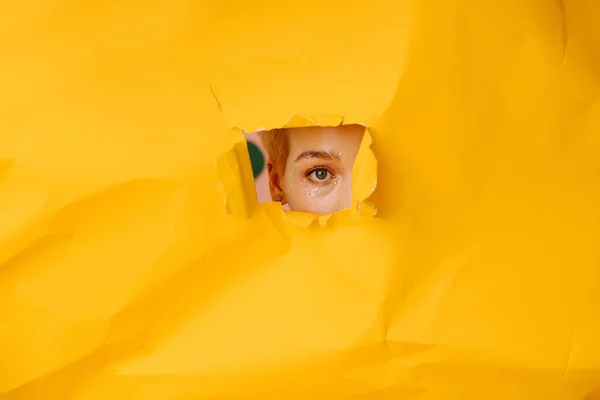 Young woman peeking with one eye through a hole in a big sheet of cramped yellow paper. She has short dyed blond hair and stars eye makeup.