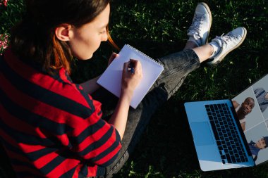 Not defterini pastel boyayla çizen mutlu genç bir kadın dizüstü bilgisayardaki video konferansından talimatlar alıyor. Yumuşak bahar güneşinin altında çimenlerde oturuyor..