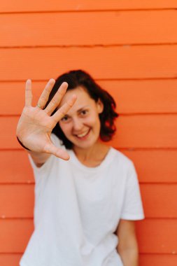 Turuncu ev duvarına karşı beyaz tişörtlü, kendinden emin gülümseyen esmer kadının yaz portresi. Kameraya el sallamak..