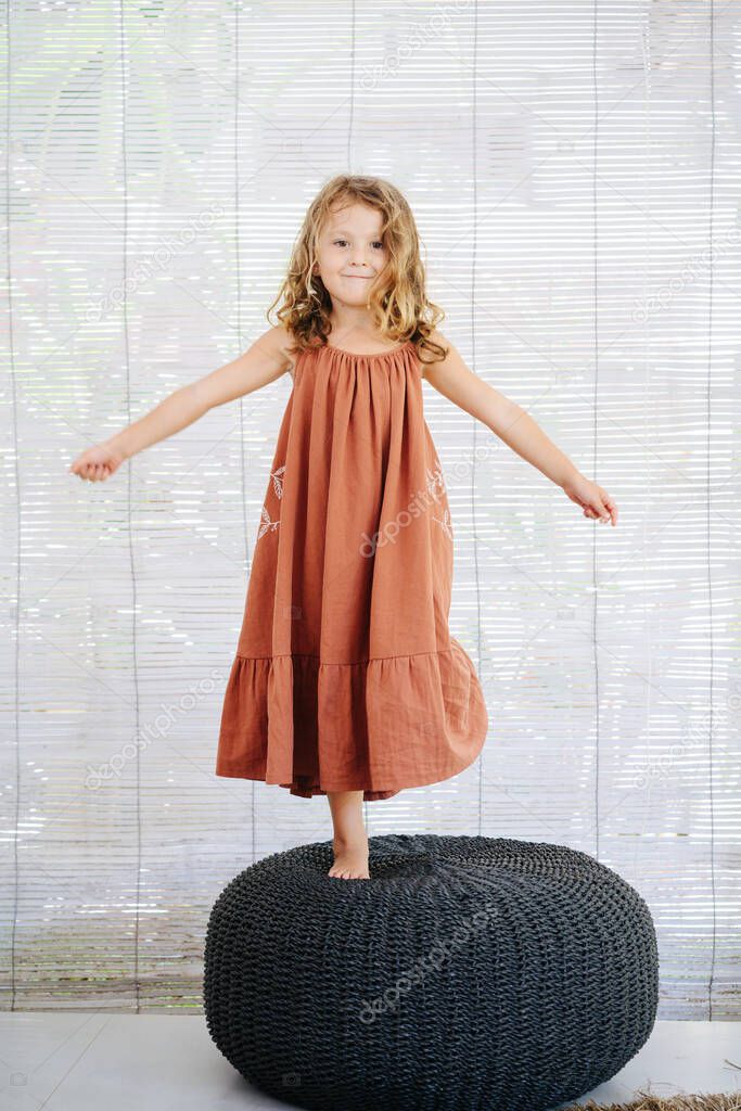 Happy little girl in long dark orange dress standing on a high black wicker pillow, trying to maintain balance. Looking at camera.