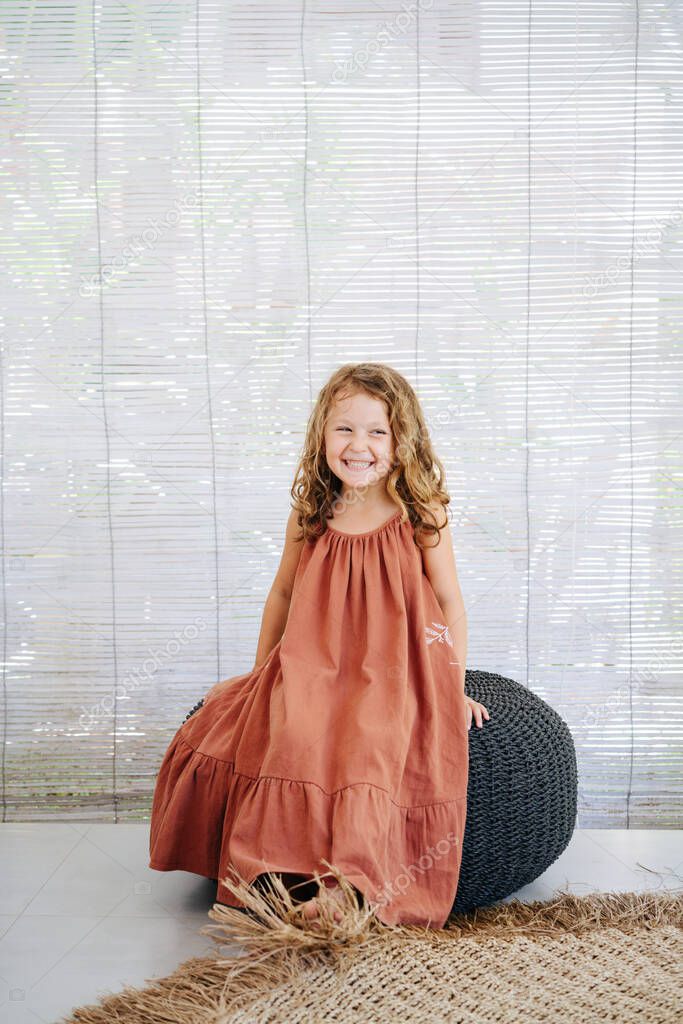 Cheerful happy little girl in long dark orange dress sitting on a high black wicker pillow, posing for a photo. Looking to side, laughing