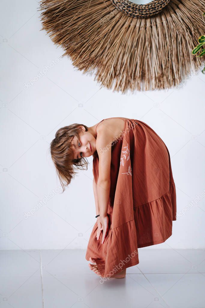 Cute barefoot little girl in a maxi dark orange dress, posing for a photo in a tropical style room. She's standing sideways on her heels, bending, reaching for her toes. Looking at camera.