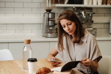 Kafede masanın arkasında çalışan, ciddi yüzlü olgun bir kadın. Ay çöreği, kahve ve cam su şişesi masanın üstünde..